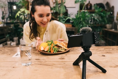 Image of a woman with a plate of food at a table, sitting facing a camera that has been set up on a small tripod