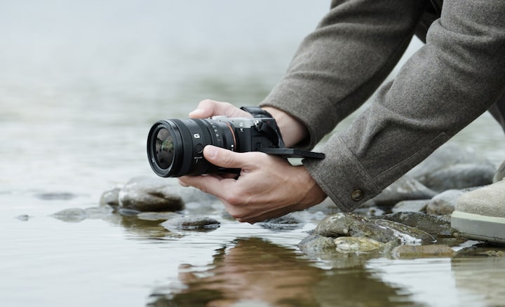 A usage image showing a user holding the α7CII with the FE 24–50mm F2.8 G near the surface of a river. The user is shooting from a low position