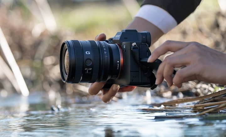 A usage image showing a user holding the α7 IV with the FE 16–25mm F2.8 G near the surface of a river. The user is shooting from a low position